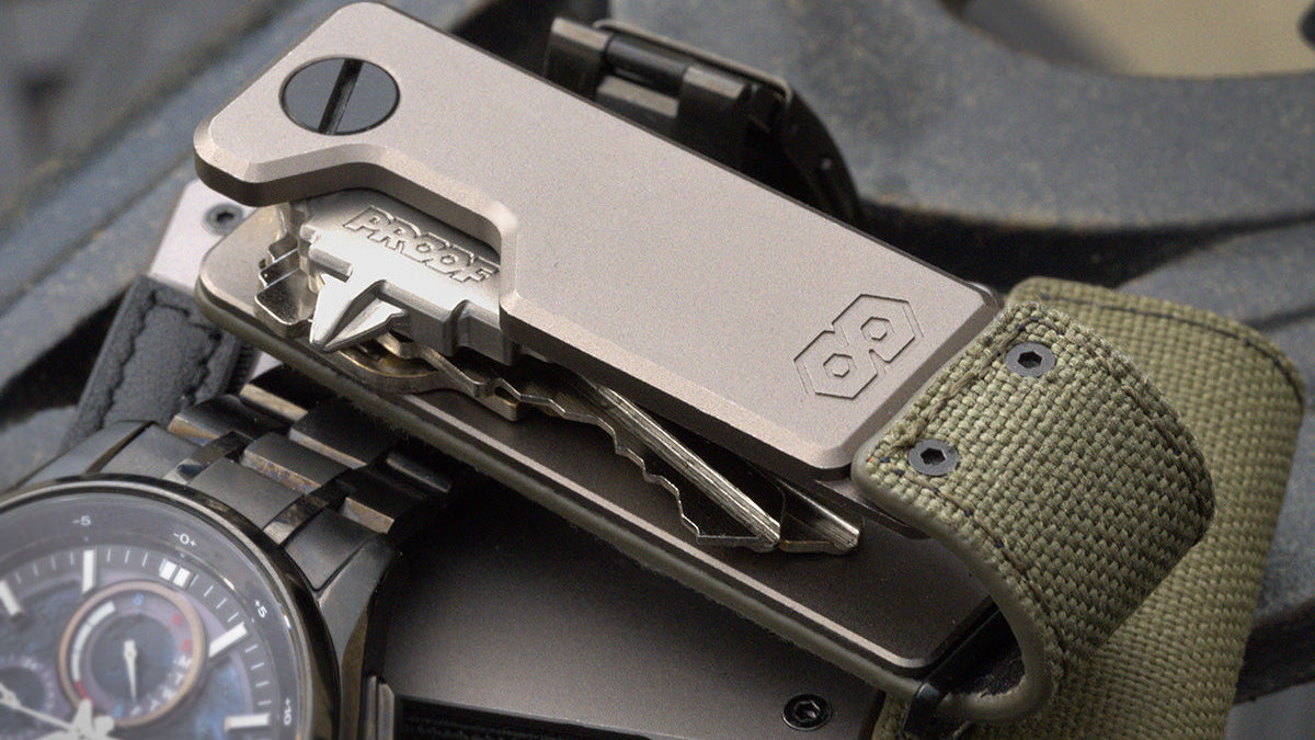Titanium PROOF key organizer with folded keys displayed open, resting on olive canvas wallet beside a Citizen Attesa chronogr