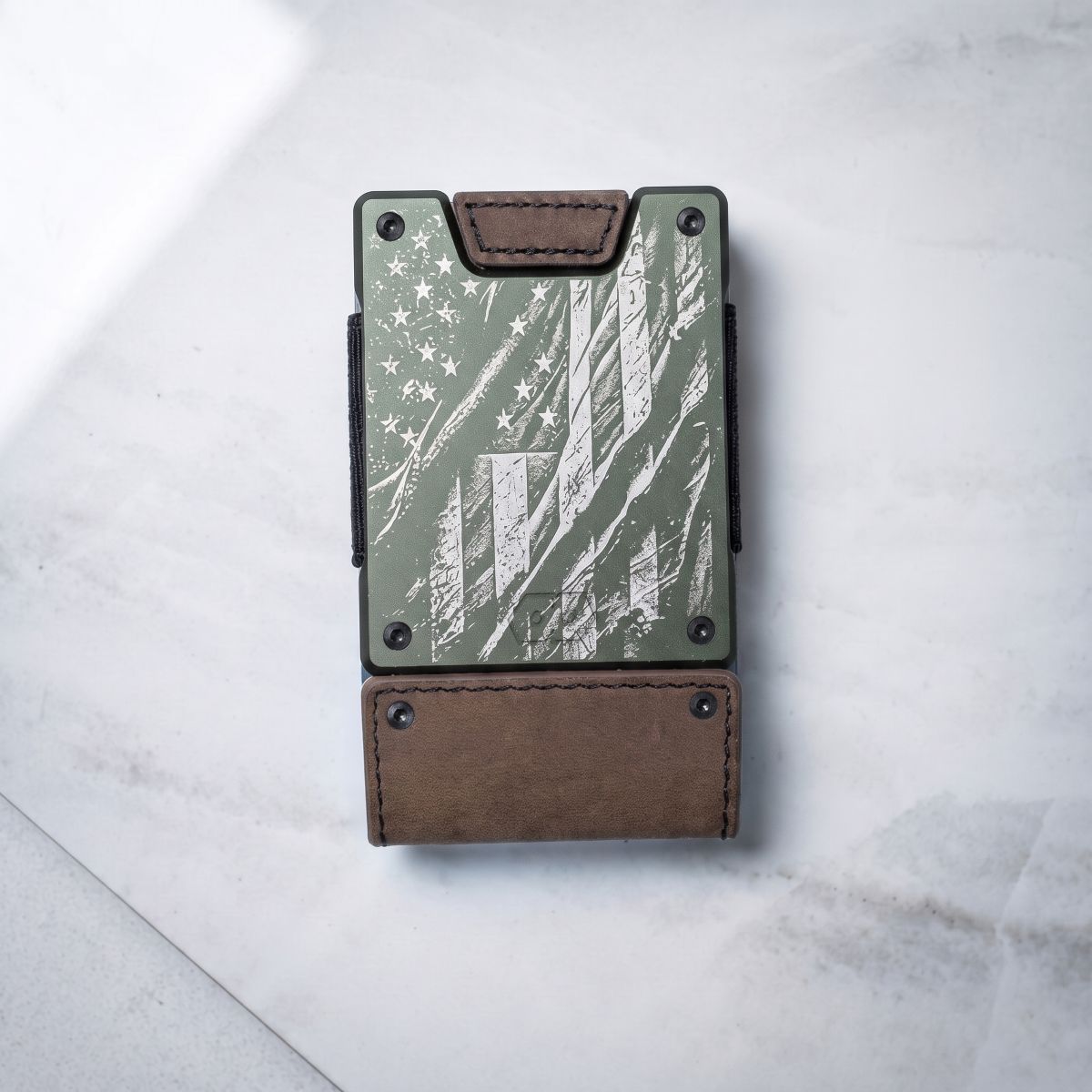 A slim black leather card holder wallet with a distressed American flag design on the front, featuring silver rivets and a black leather strap, photographed on a light gray marble surface.