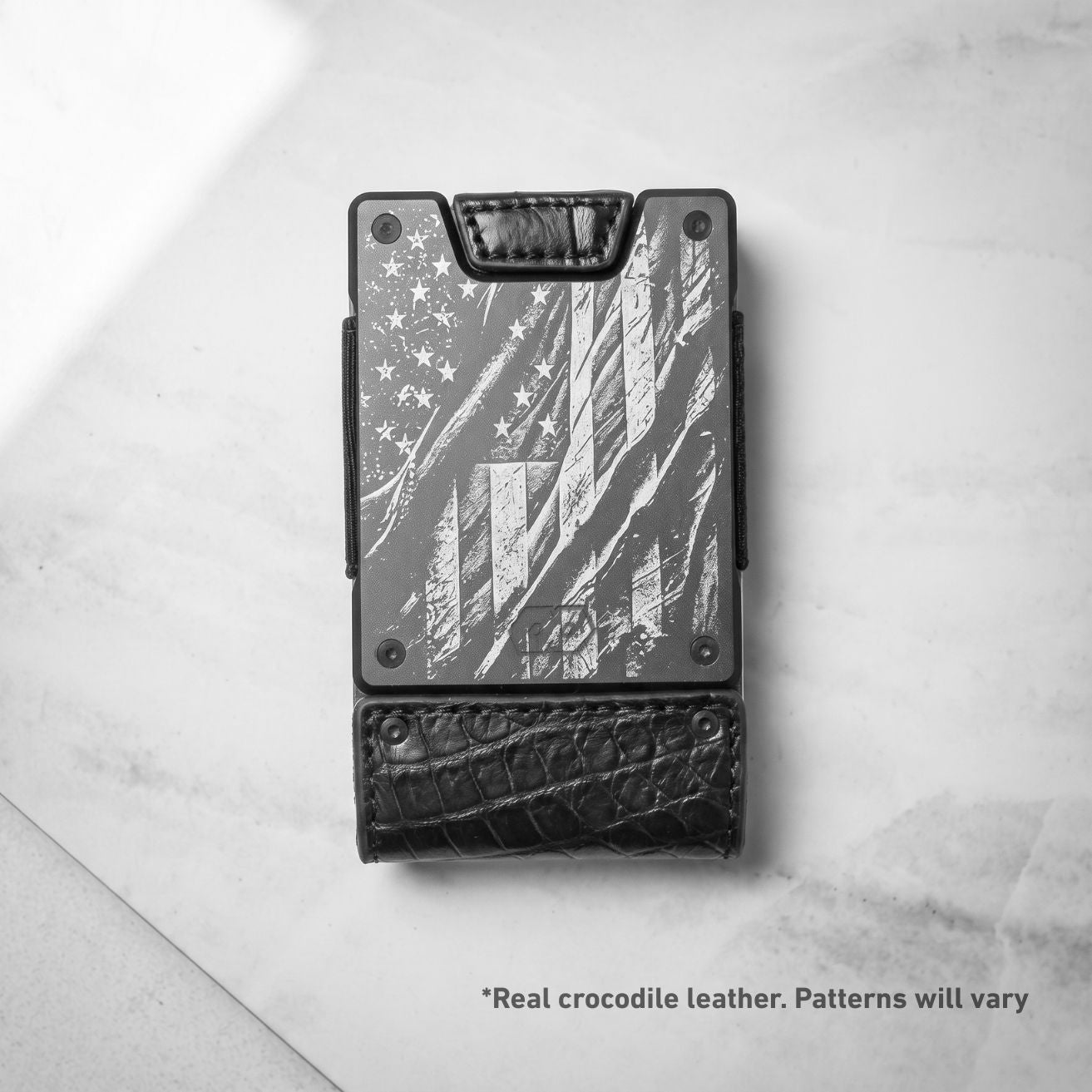 A black and gray slim wallet featuring an engraved distressed American flag design on the front and a textured black leather bottom section, photographed from above on a light marble surface.