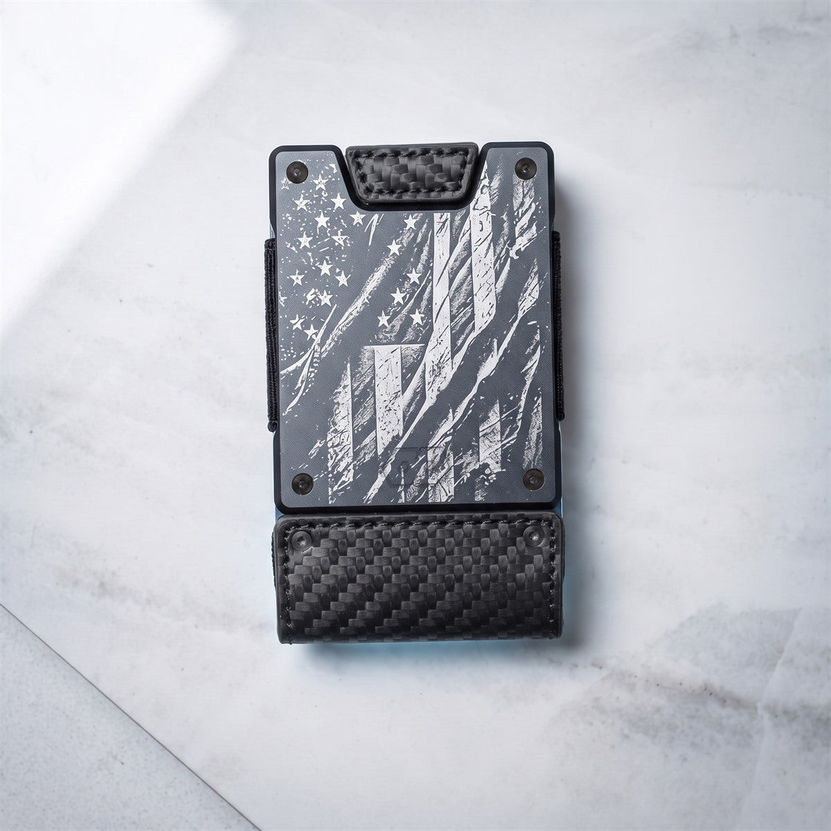 A slim black leather card holder wallet with a distressed American flag design on the front, featuring silver rivets and a black leather strap, photographed on a light gray marble surface.