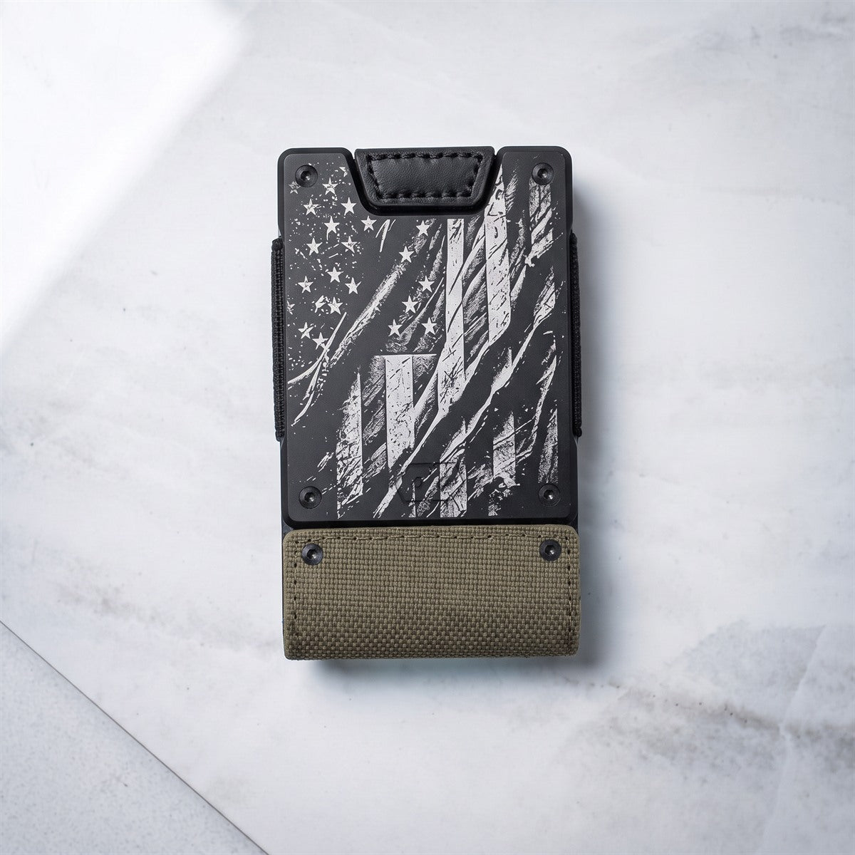 A slim black and olive drab wallet with a distressed American flag design, photographed flat on a light gray marble surface.
