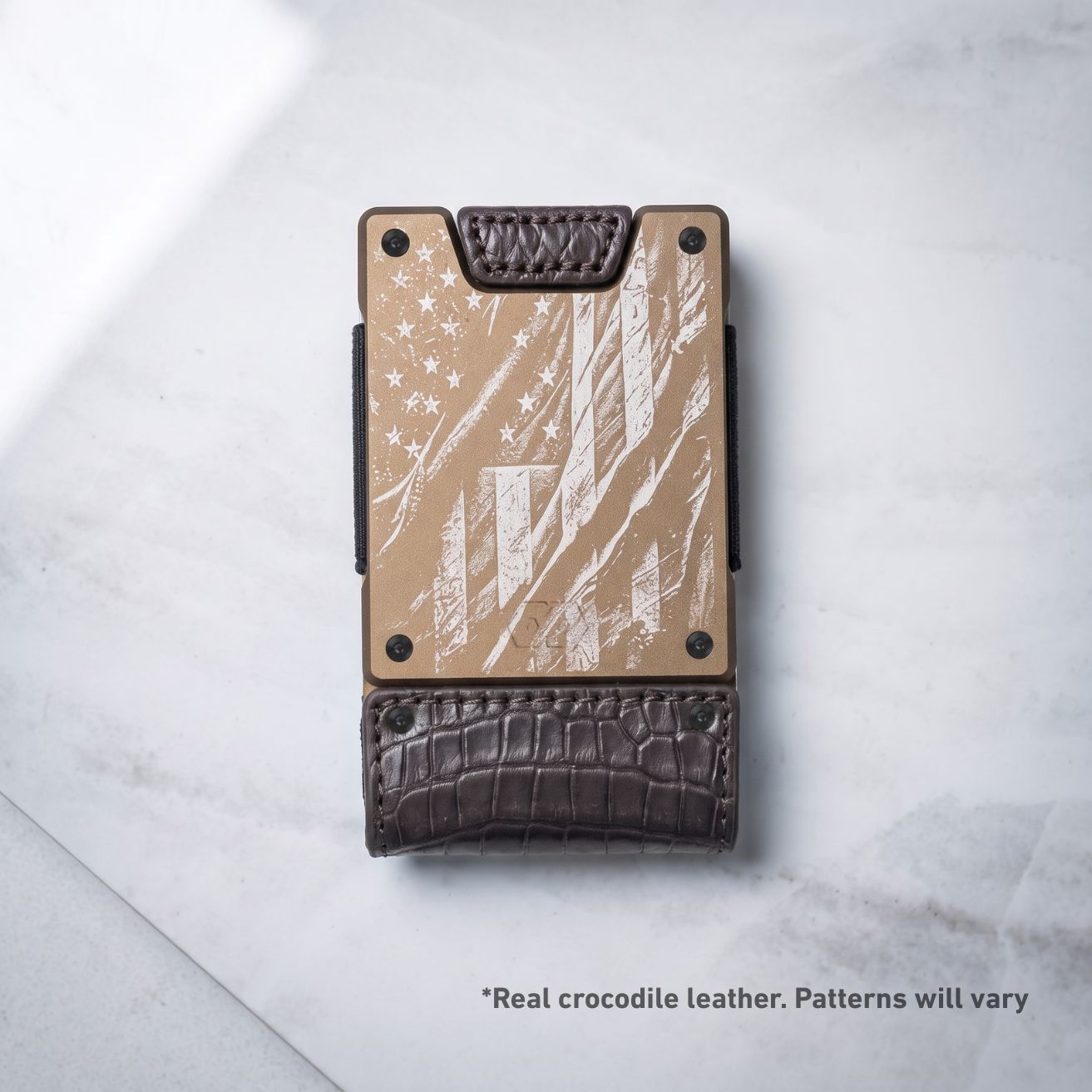 A minimalist metal wallet with an etched American flag design on a tan background, finished with a dark brown crocodile-embossed leather base and top loop, photographed on a light marble surface.