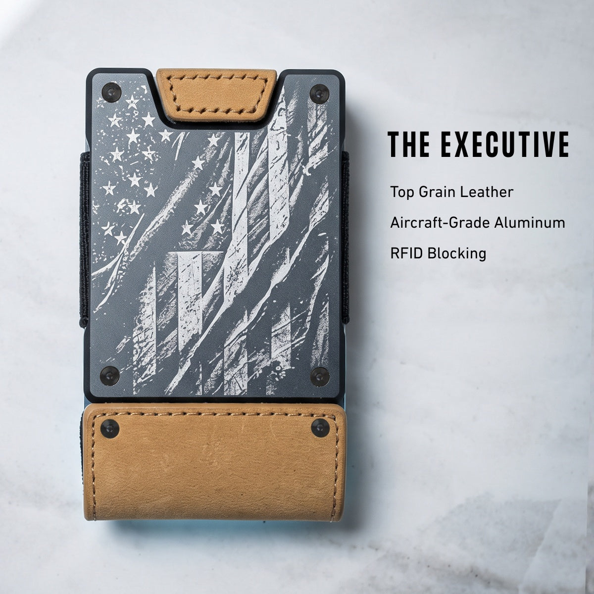 A slim wallet card holder with a dark gray and white distressed American flag design, tan leather trim, and a tan leather bottom loop, shown on a light marble background with text reading 'THE EXECUTIVE' and product features.