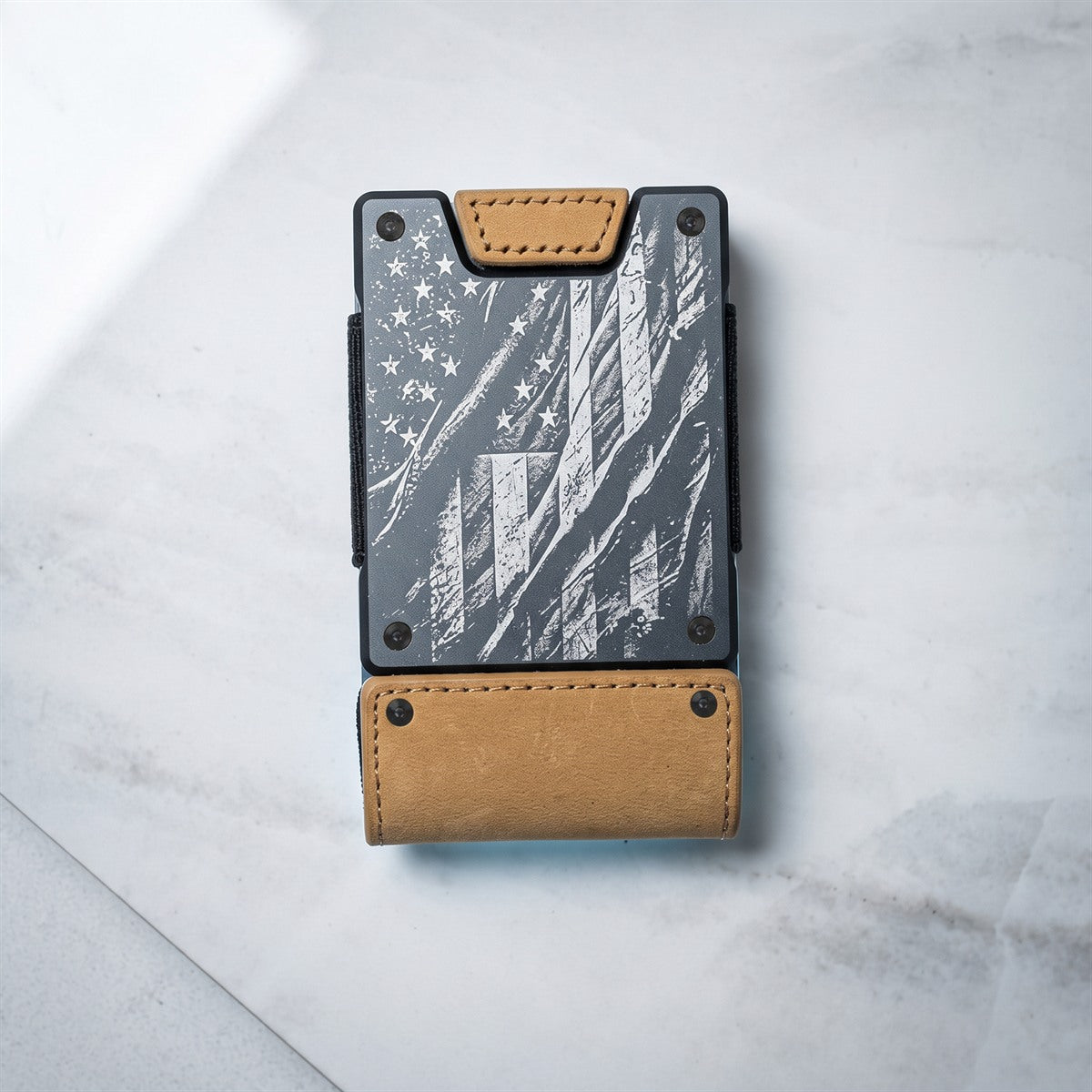 A minimalist card holder wallet with a black and gray distressed American flag design and tan leather accents, photographed from above on a light gray marble surface.
