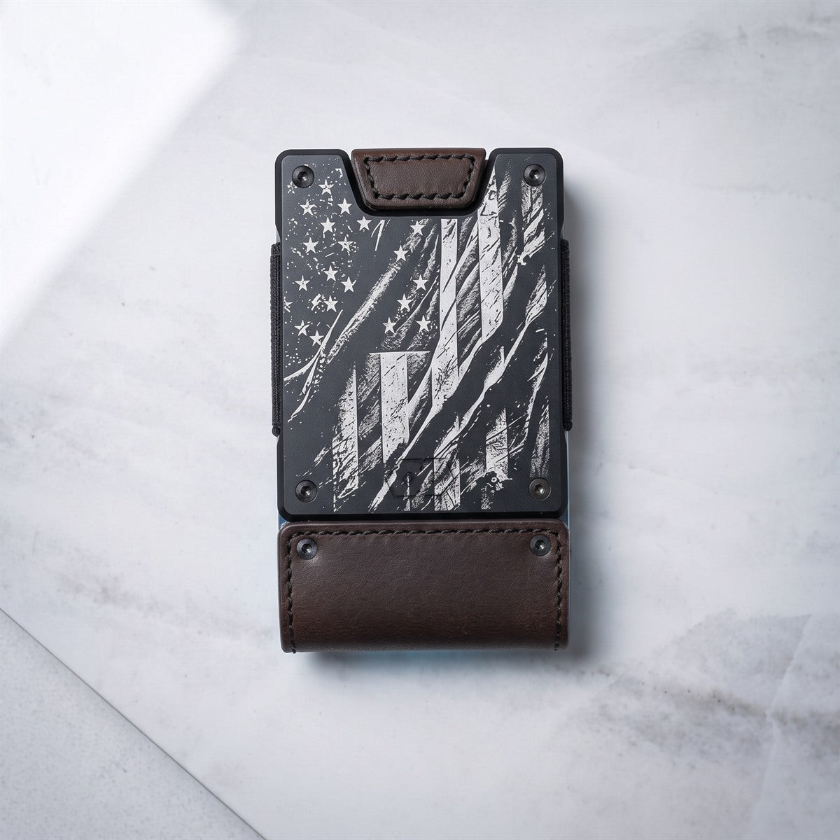 A slim black and brown wallet with a distressed American flag design on the front, resting on a light gray marble surface.