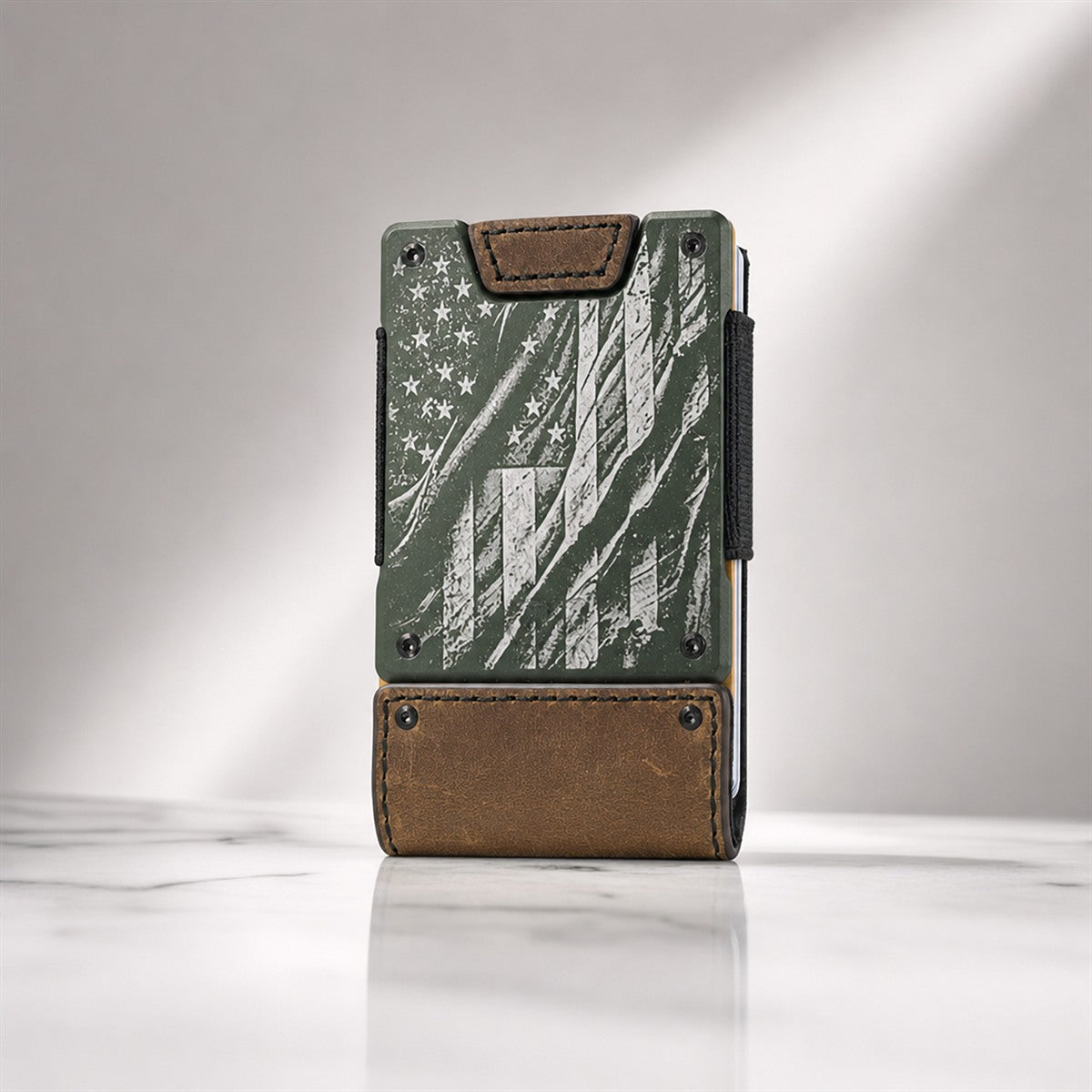 A slim vertical wallet or card holder standing upright on a light marble-like surface against a soft gray studio background. The product has a distressed American flag design in muted green, brown, and white, with brown leather-like trim at the top and bottom and a black side strap.