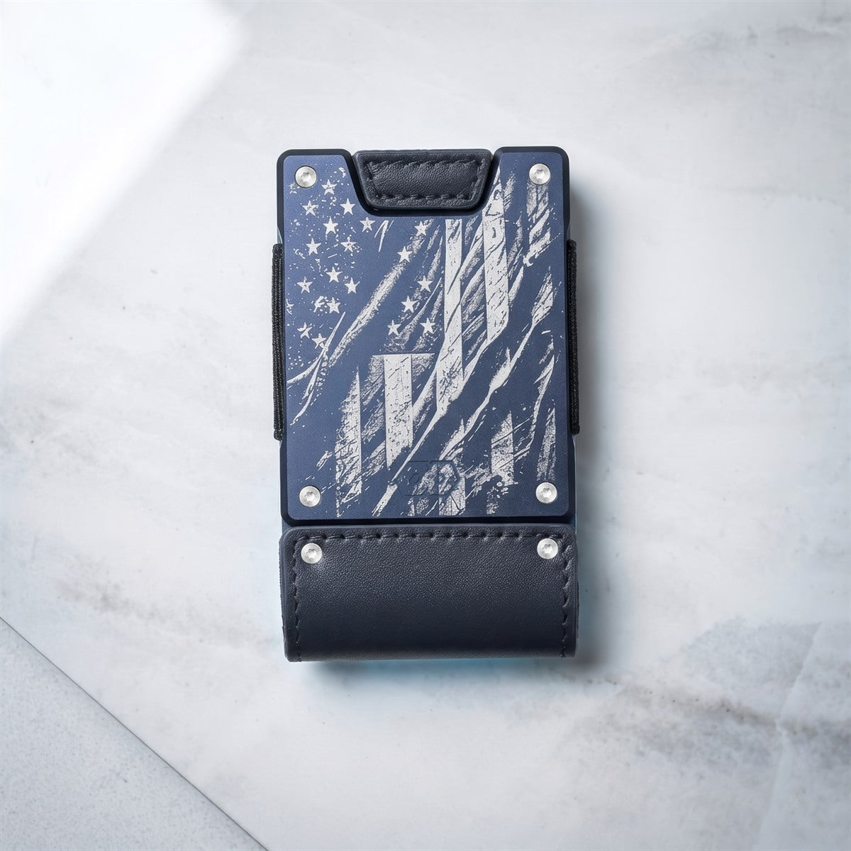 A slim black leather card holder wallet with a distressed American flag design on the front, featuring silver rivets and a black leather strap, photographed on a light gray marble surface.