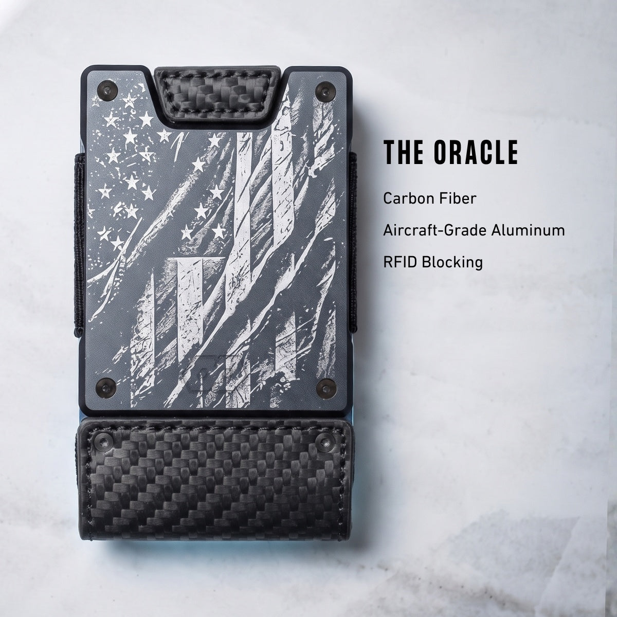 A slim black and gray wallet shown from above on a light marble-like surface, with text to the right reading 'THE ORACLE' and material callouts: Carbon Fiber, Aircraft-Grade Aluminum, RFID Blocking. The wallet has a distressed American flag graphic on the front and a carbon-fiber-look lower section.