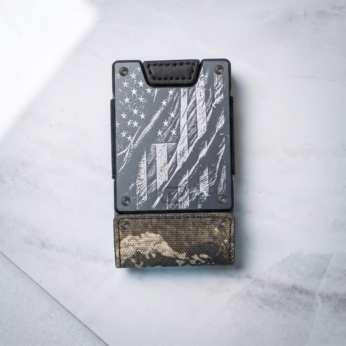 A slim black wallet with a distressed American flag design on the front and a camouflage-patterned lower section, photographed from above on a light marble surface.